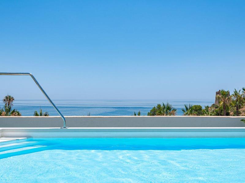Uitzicht op een zwembad met helderblauw water en palmbomen met de zee op de achtergrond.