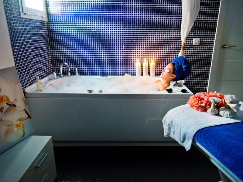 Vrouw ontspant in bruisend bad met kaarsen voor blauw betegelde wand.