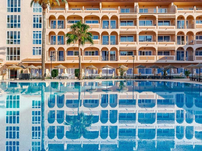 Groot zwembad voor een meerlaags hotel met palmbomen en blauwe parasols.