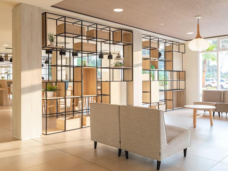 Ampia hall d'hotel con divisorio metallico, poltrone grigie e luce naturale.