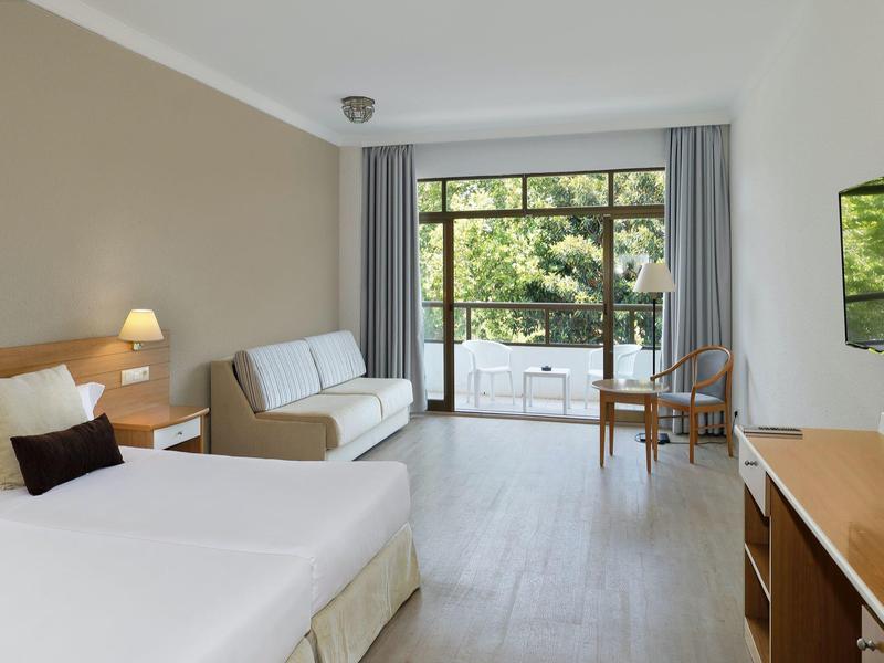 Camera d'hotel luminosa con letto matrimoniale, divano, balcone e TV a parete.