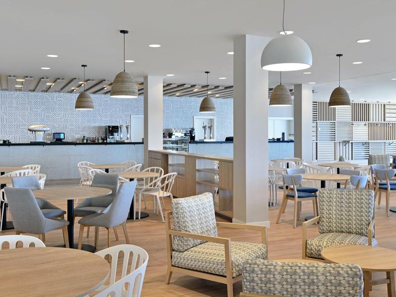 Ampia sala d'hotel moderna con sedie, tavoli rotondi e molta luce naturale.