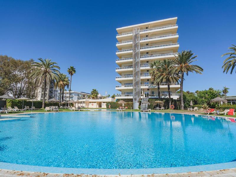 Grande piscina rotonda davanti a un hotel moderno con palme e cielo blu.