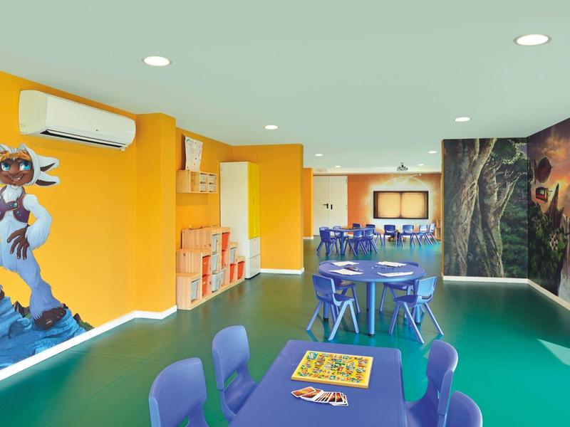 Sala giochi colorata con tavoli, sedie blu e decorazioni murali animate.