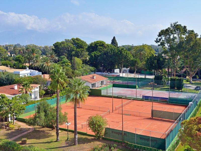 Campi da tennis in terra rossa circondati da alberi e edifici sotto un cielo sereno.