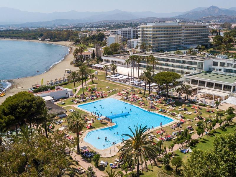 Vista aerea di un complesso alberghiero con piscina vicino a una spiaggia con palme.