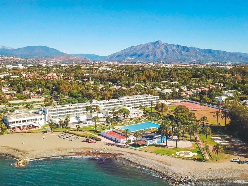 Hotel sul mare con giardini, piscina, spiaggia e montagne sullo sfondo.