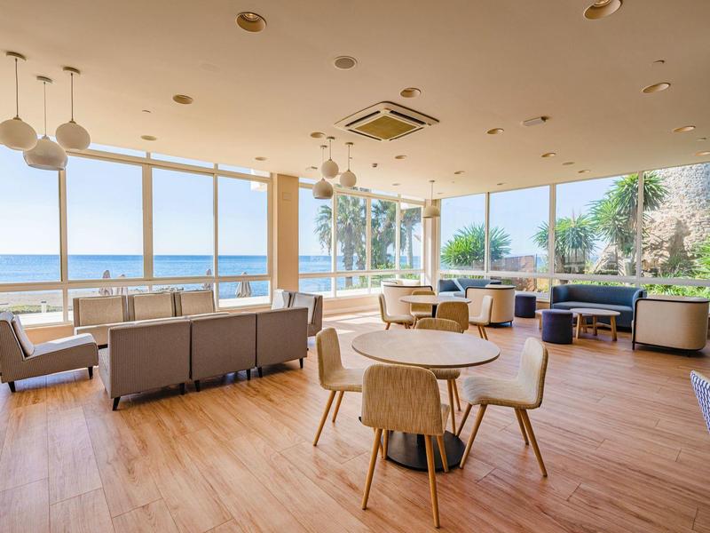 Sala luminosa con grandi finestre sul mare, tavoli e sedie moderne.