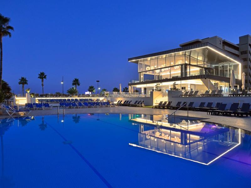 Moderní hotel s osvětleným bazénem a palmami za soumraku.