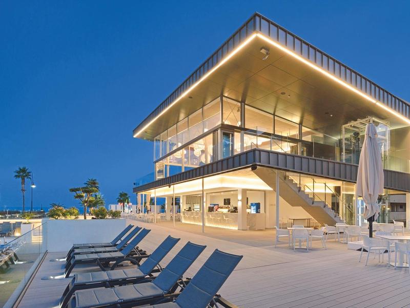 Moderní hotel s osvětlenou terasou a lehátky u bazénu večer.