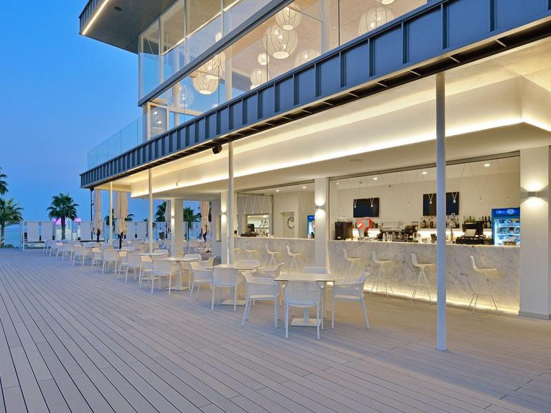 Moderní bar u moře s bílými stoly a teplým osvětlením za soumraku.