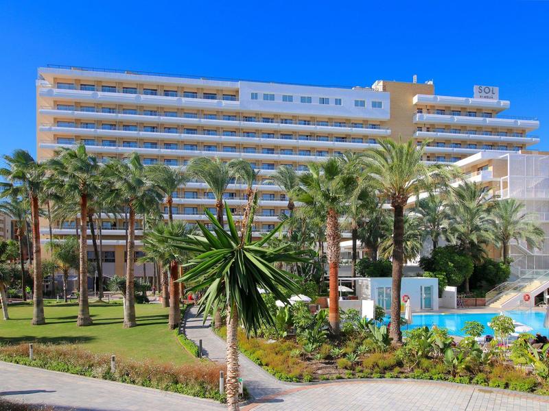 Moderní hotel s bazénem obklopeným palmami pod jasnou modrou oblohou.