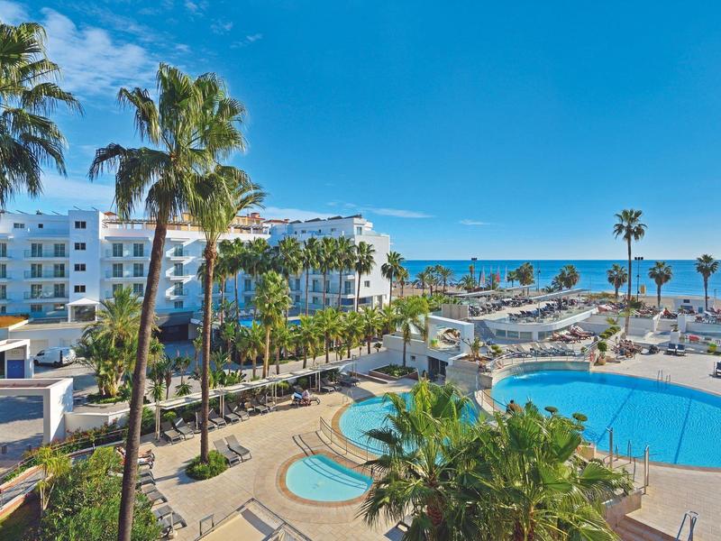 Pohled na hotelový bazén s palmami, bílými budovami a mořem v pozadí pod modrou oblohou.