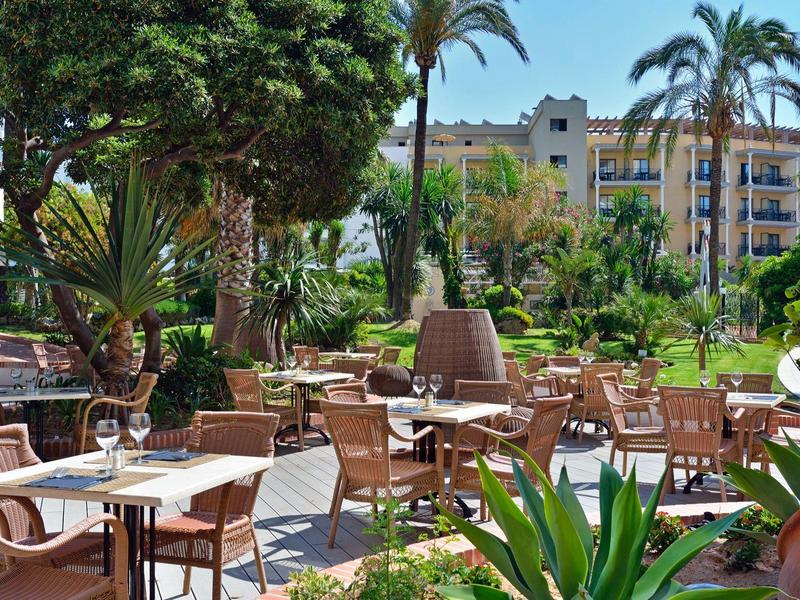 Venkovní terasa s stolky a židlemi, obklopená palmami a bujnou zelení.