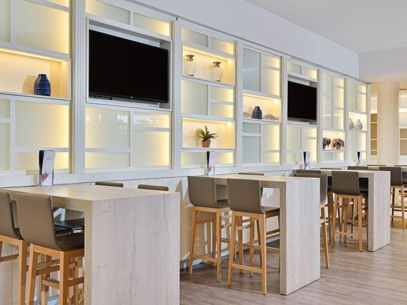 Světlá místnost s vysokými stoly, dřevěnými stoličkami a osvětlenými policemi.