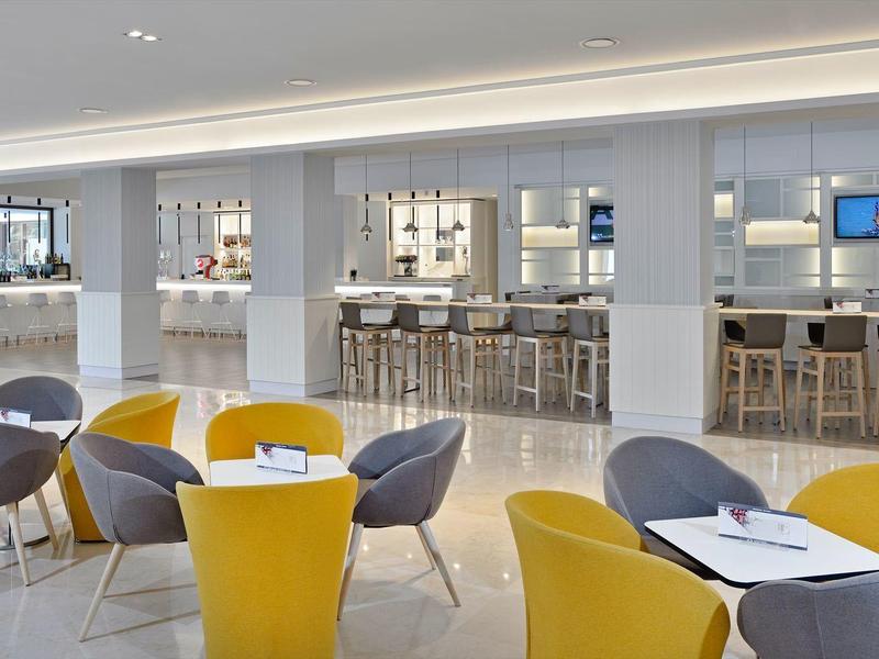 Světlý salonek se žlutými a šedými křesly kolem malých bílých stolů.