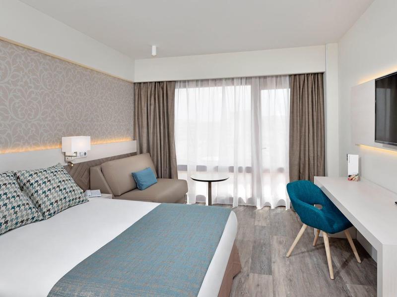 Moderní hotelový pokoj s manželskou postelí, pohovkou, psacím stolem a světlým balkonem.