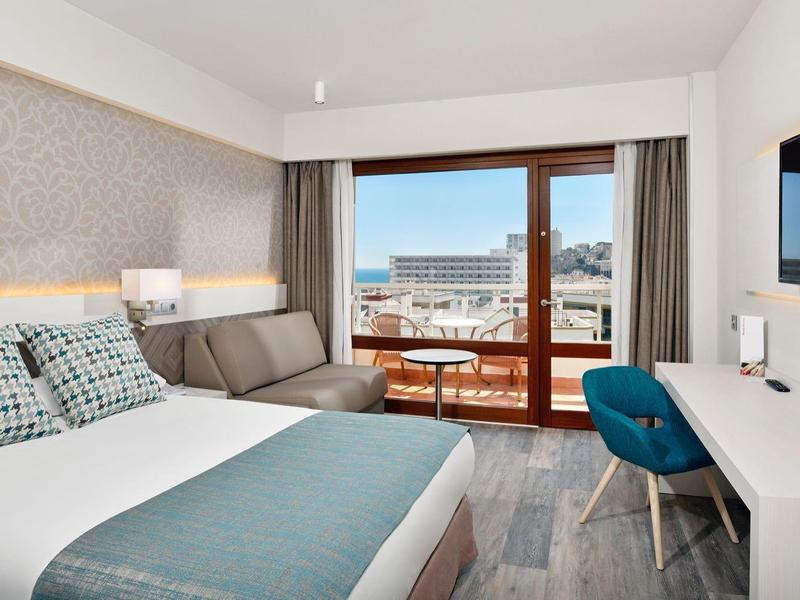 Moderní hotelový pokoj s manželskou postelí, psacím stolem, křeslem a balkonem s výhledem na moře.