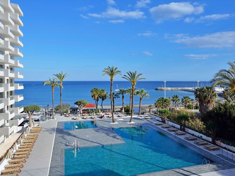 Piscine d'hôtel près de la mer avec palmiers et ciel bleu clair.