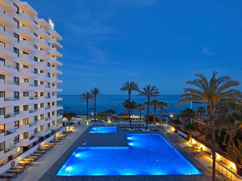 Hôtel avec grande piscine éclairée, palmiers, vue sur la mer au crépuscule.