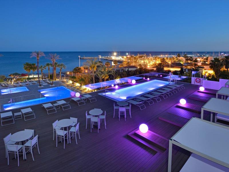 Piscine éclairée en violet avec vue sur la mer et tables blanches au bord du toit la nuit.