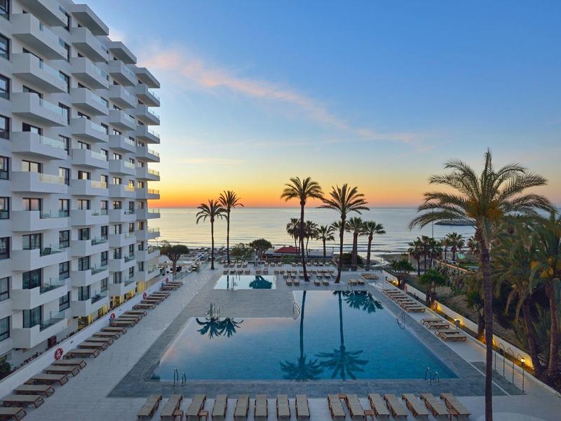 Piscine avec chaises longues près de la mer au coucher du soleil avec un grand bâtiment blanc.