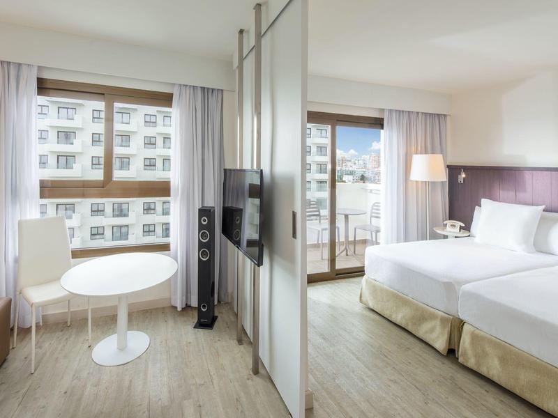 Chambre d'hôtel moderne avec lits jumeaux, table, chaises, et télévision suspendue.