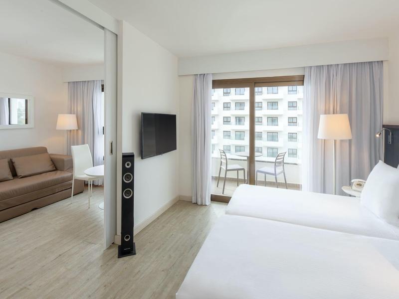 Chambre d'hôtel moderne avec lit double, canapé, TV et balcon lumineux.