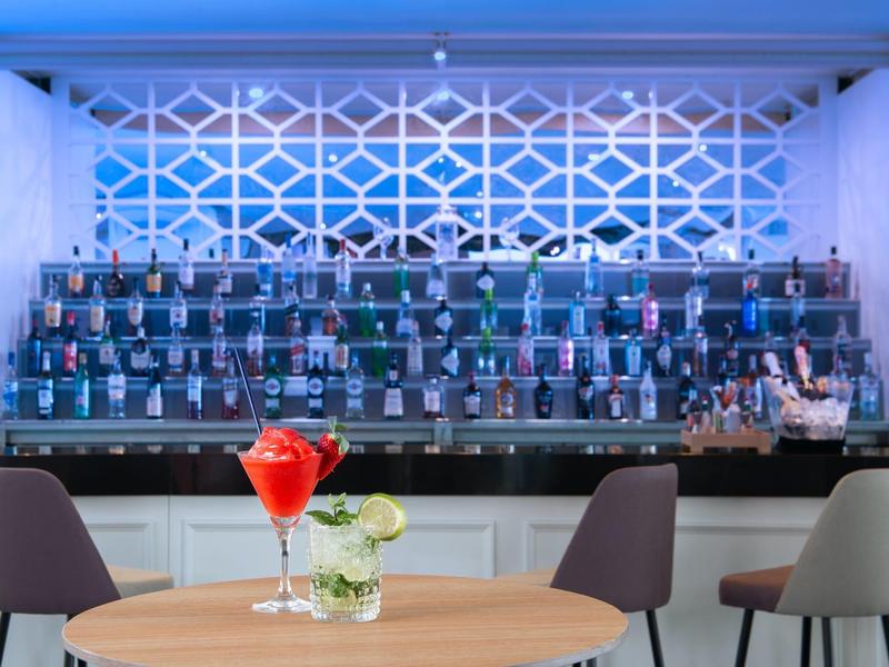 Bar moderne avec bouteilles illuminées et table avec cocktails rouges et fleurs blanches.