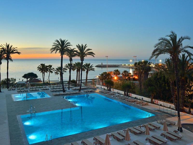 Piscine éclairée au coucher du soleil avec palmiers et vue sur la mer.