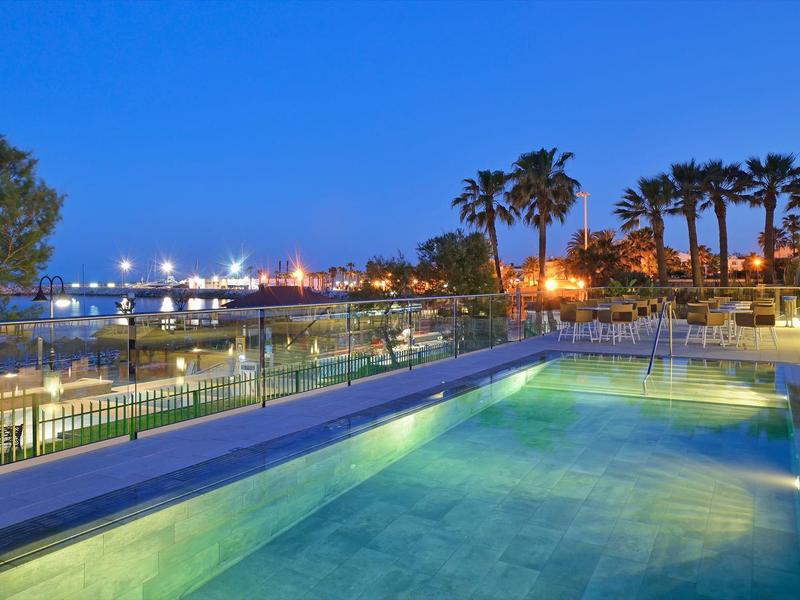 Piscine éclairée au bord de mer avec palmiers et ciel bleu nocturne.