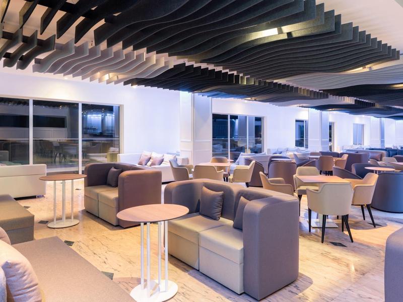 Salon d'hôtel moderne avec canapés gris, tables et grands stores sur les fenêtres.