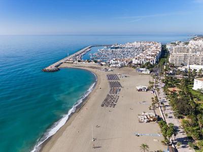 Luchtfoto van een strand met ligbedden, haven en nabijgelegen gebouwen onder heldere lucht.