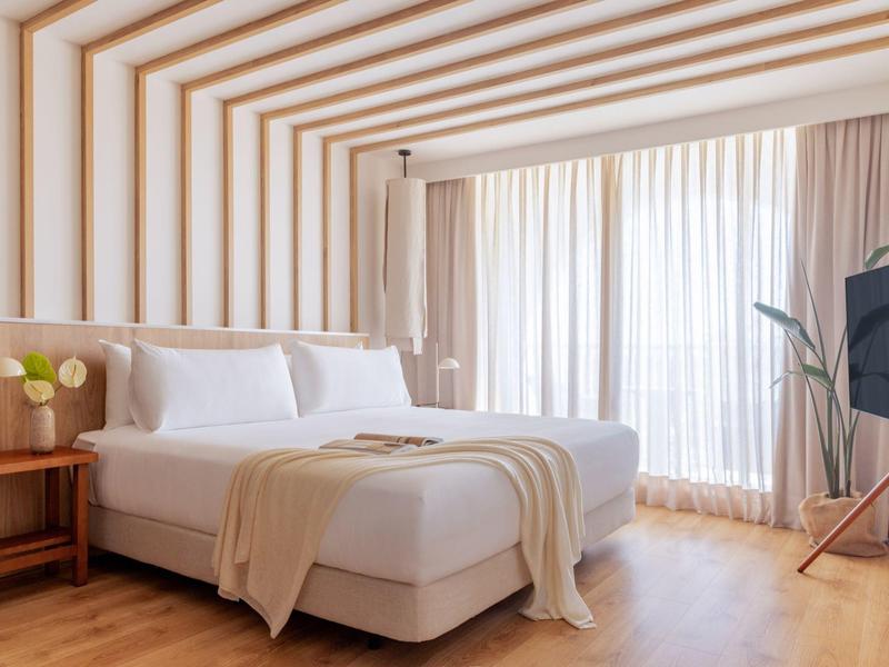 Modern hotelkamer met groot bed, houten vloer en raam met doorschijnende gordijnen.