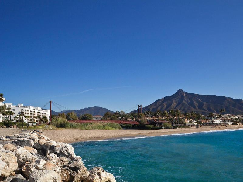 Strandzicht met rotsen, blauwe zee, zand en bergen op de achtergrond onder een heldere lucht.