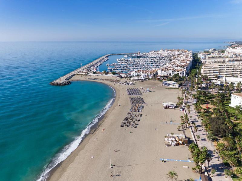 Luchtfoto van een strand met ligbedden, haven en nabijgelegen gebouwen onder heldere lucht.