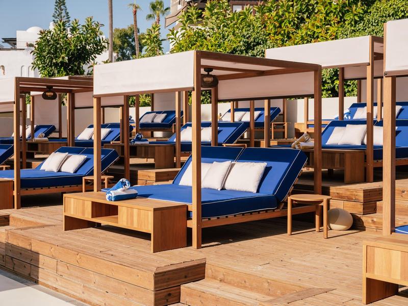 Luxe strandbedden met blauwe kussens en houten tafels op een houten buitendek.