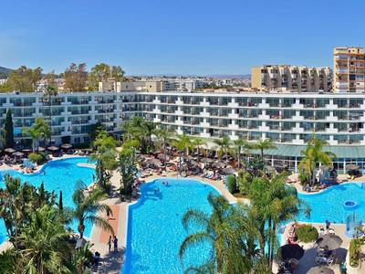 Velký hotel s několika bazény, slunečními parasoly a palmami za slunečného dne.