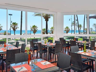 Moderní restaurace s výhledem na moře, velkými okny a tmavými židlemi.