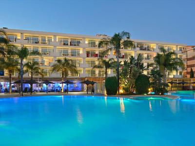 Hell erleuchtetes Hotel mit Palmen und großem Pool im Außenbereich bei Sonnenuntergang.
