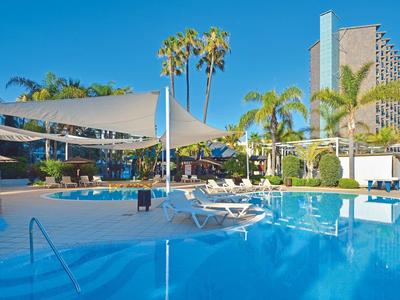 Velký hotelový bazén s lehátky a plachtami, obklopený palmami a vysokými budovami.