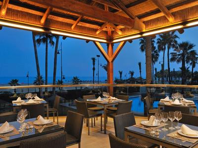 Überdachte Terrasse eines Restaurants mit Tischen und Blick auf Palmen und den Pool bei Dämmerung.