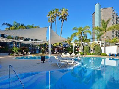 Hotelový bazén s lehátky, stínícími plachtami a palmami pod jasnou oblohou.