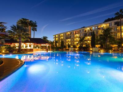 Osvětlený hotelový bazén v noci s přilehlou budovou a palmami.