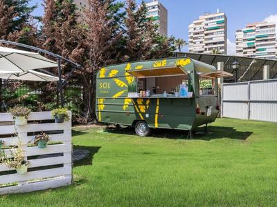 Grüner Food-Truck auf Rasenfläche neben weißen Gartenmöbeln und Gebäuden im Hintergrund.