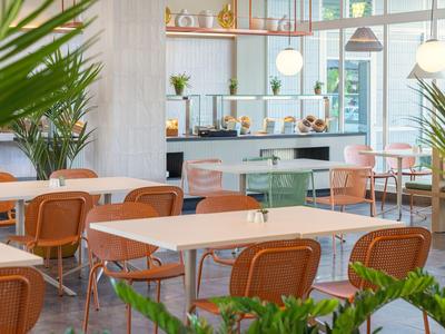 Moderner Speisesaal mit weißen Tischen, orangefarbenen Stühlen und Pflanzen im Hintergrund.