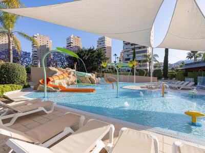 Moderner Hotelpool mit Liegestühlen und Sonnensegeln unter blauem Himmel, umgeben von Gebäuden.