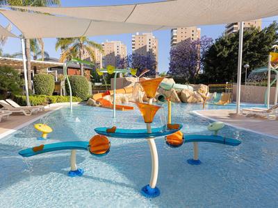 Bunter Wasserspielplatz mit Rutschen und Wasserfontänen in einem Hotelpool bei sonnigem Wetter.