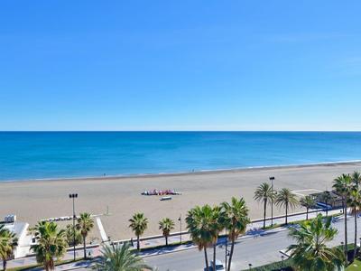 Weiter Sandstrand mit Palmen und einer Straße am Ufer unter klarem blauem Himmel.