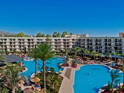 Velký hotel s bazénem a mnoha lehátky pod modrou oblohou.